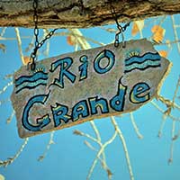 Photograph of painted sign reading Rio Grande hanging from branch.